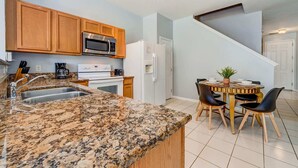A kitchen with granite countertops, wooden cabinets, a stainless steel microwave, a white refrigerator, and a dining area with a wooden table and chairs. - Relaxing 3BR townhome w/ resort access (Kissimmee)