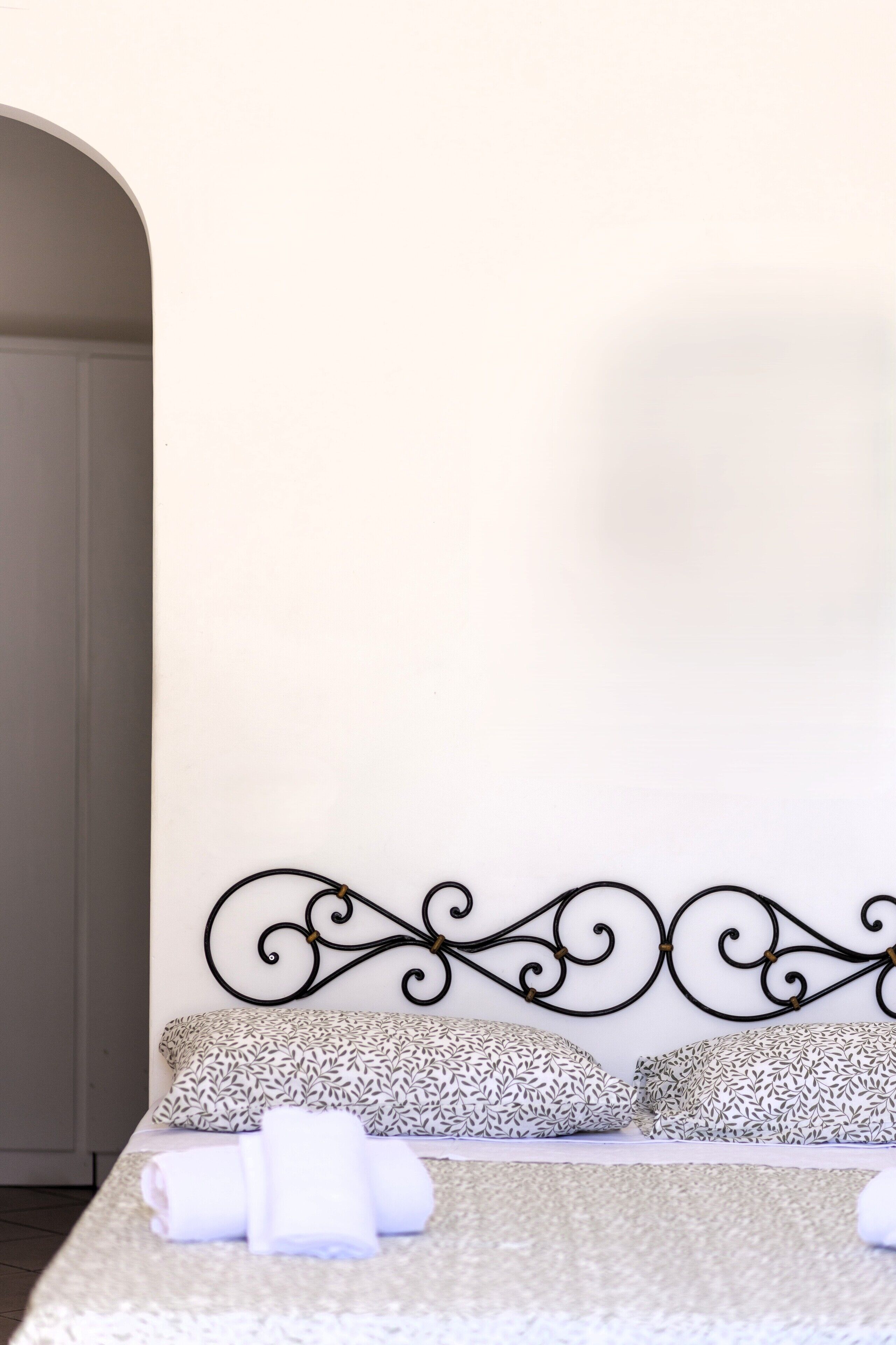 A neatly made bed with patterned pillows and a rolled towel, a decorative metal headboard, and a plain wall.