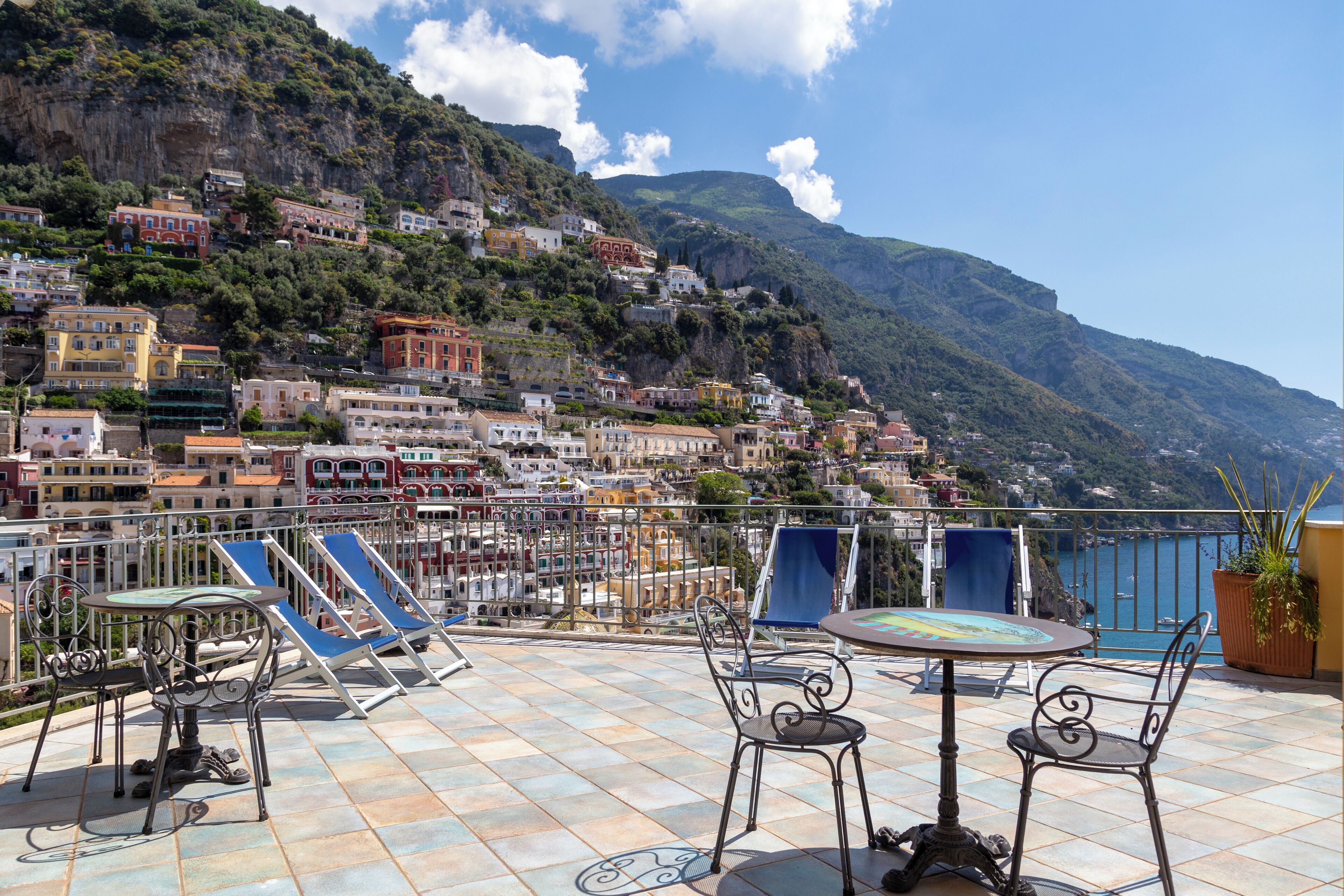 A terrace with outdoor seating, a view of a hillside town, and a body of water.