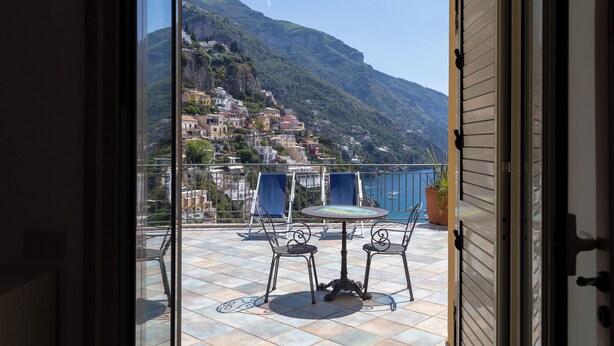 A balcony with a table and chairs, overlooking a coastal town with buildings on a hillside. - Sea Soul Positano (Positano)