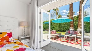 Un dormitorio con una cama grande, una mesita de noche, una lámpara y un ventanal corredizo que da a un patio exterior con un sofá, una mesa y sombrillas.