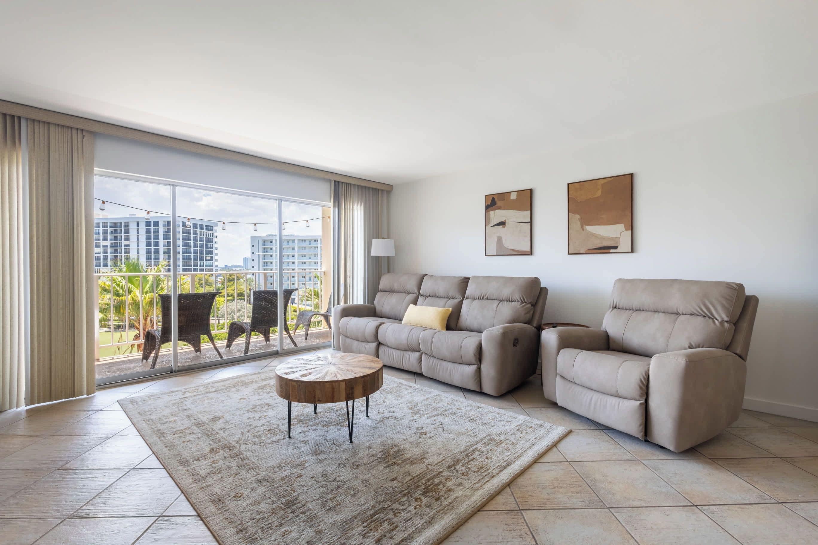 A spacious living room with a large sliding glass door leading to a balcony, two sofas, a coffee table, and two framed artworks on the wall.