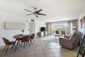 A spacious living area with a dining table, chairs, a sofa, a TV, and a balcony with a city view.