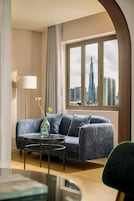 A modern living room with a grey sofa, a round coffee table, and a floor lamp. A large window offers a view of a city skyline.