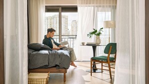 Un dormitorio moderno con cama, un sillón de lectura, una mesita de noche con lámpara y vistas a edificios altos.