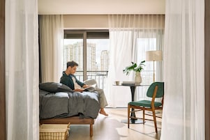 A modern bedroom with a bed, a reading chair, a small table with a lamp, and a view of high-rise buildings.