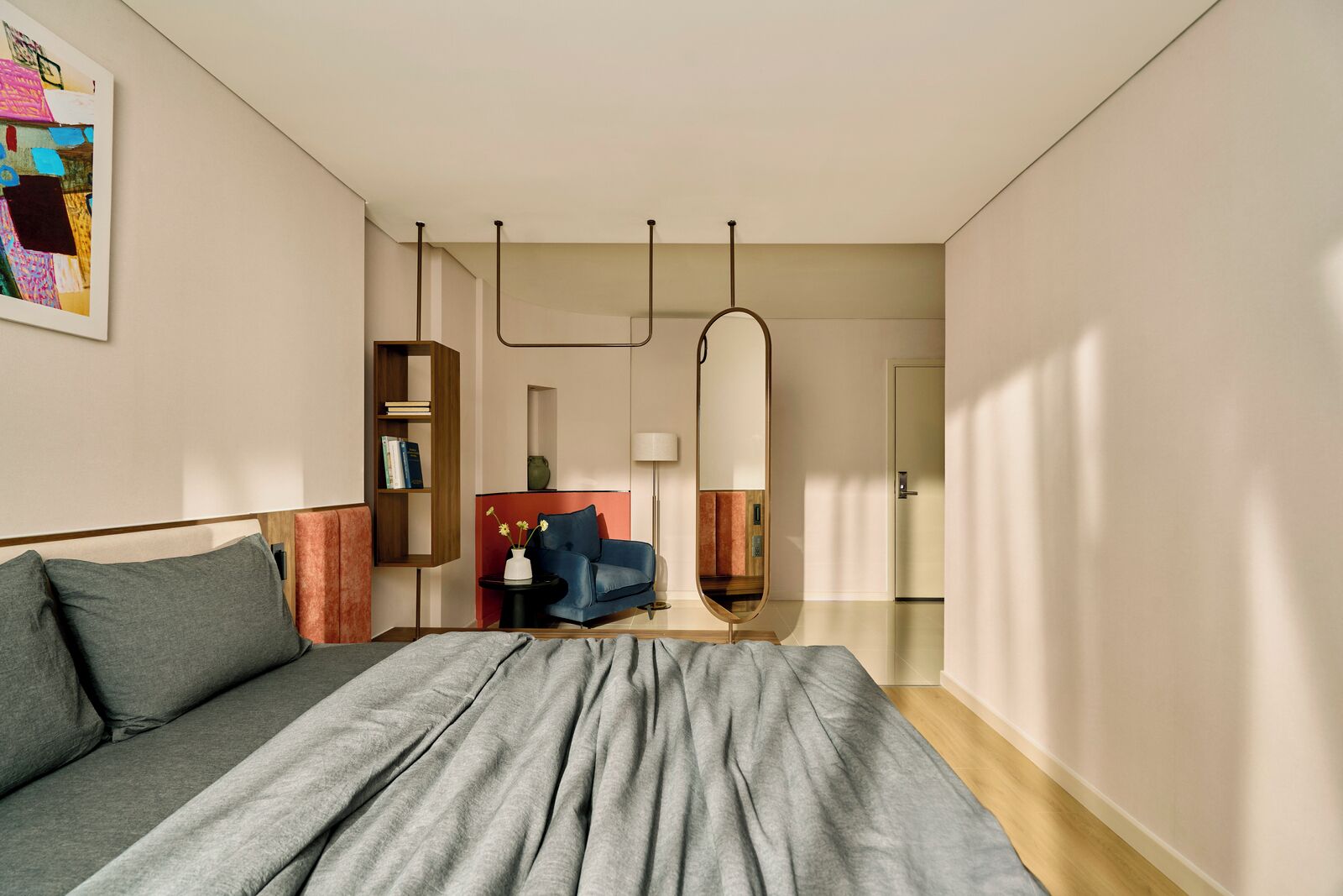 A modern bedroom with a large bed, a blue armchair, a bookshelf, and a mirror.