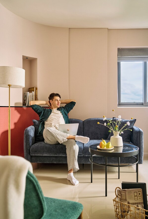 Une personne est assise sur un canapé bleu foncé. Sur une table d’appoint à côté du canapé, on aperçoit un ordinateur portable, un vase rempli de fleurs et un panier de fruits.