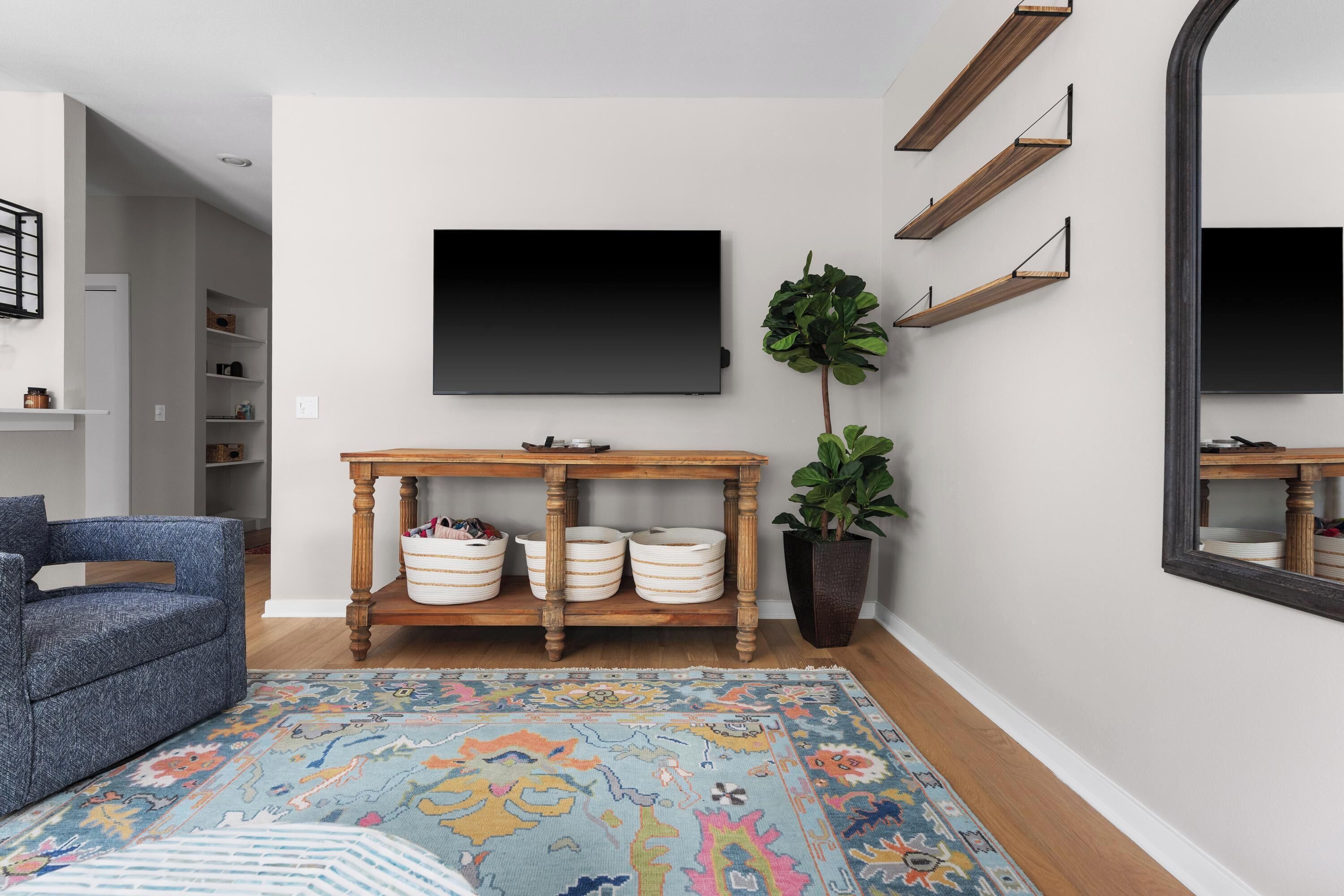 A living room with a blue sofa, a wooden coffee table with white bowls, a large potted plant, and a flat-screen TV mounted on the wall.