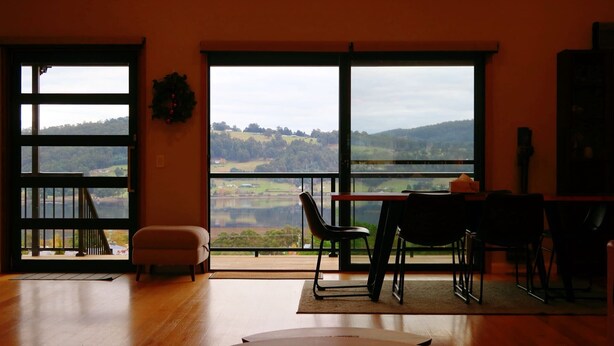 Ein Zimmer mit großen Fenstern, Holzboden, einem runden Tisch und Blick auf einen See und Hügel.