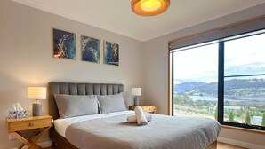 Ein Schlafzimmer mit einem großen Bett, zwei Nachttischen, einem Fenster mit Aussicht und drei an der Wand befestigten Kunstwerken.