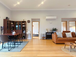 Ein geräumiges Wohnzimmer mit Holzboden, einem braunen Ledersofa, einem Esstisch mit Stühlen, einem Bücherregal und einer Klimaanlage.