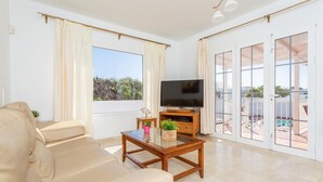 A living room with a beige sofa, a wooden coffee table with a plant, a flat-screen TV, and large windows with curtains.