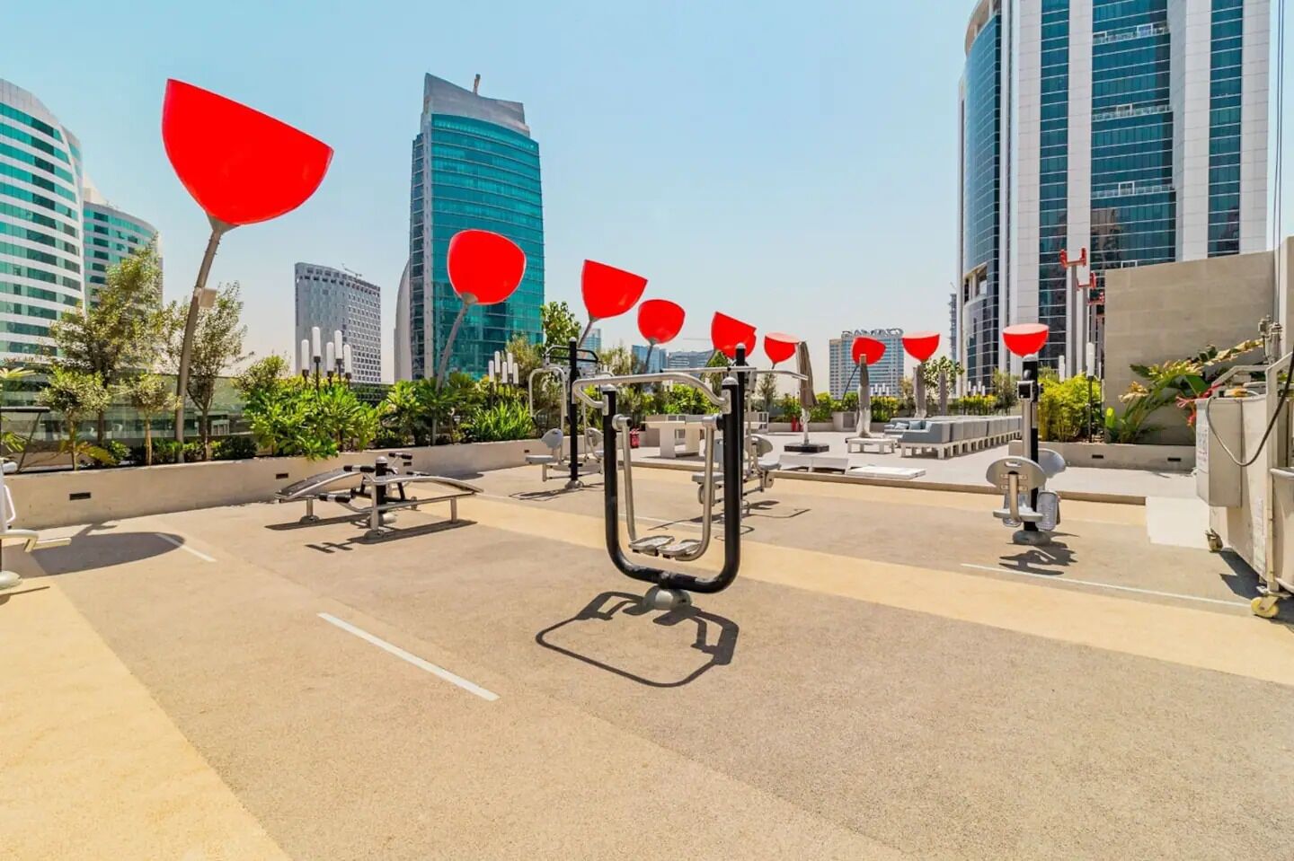 An outdoor gym area with exercise equipment and red decorative elements, surrounded by modern buildings.