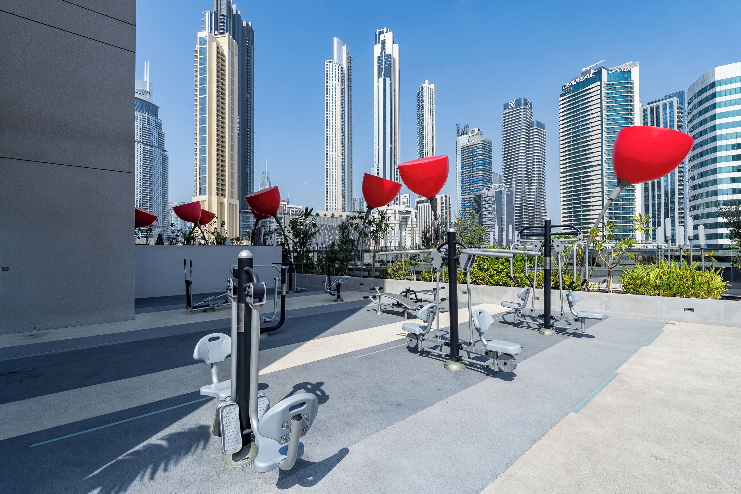 An outdoor gym with exercise equipment and red umbrellas, set against a backdrop of modern high-rise buildings.