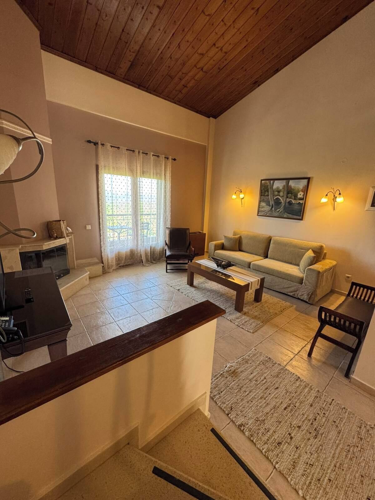A living room with a sofa, coffee table, and a TV. There is a window with curtains, a painting on the wall, and a lamp.