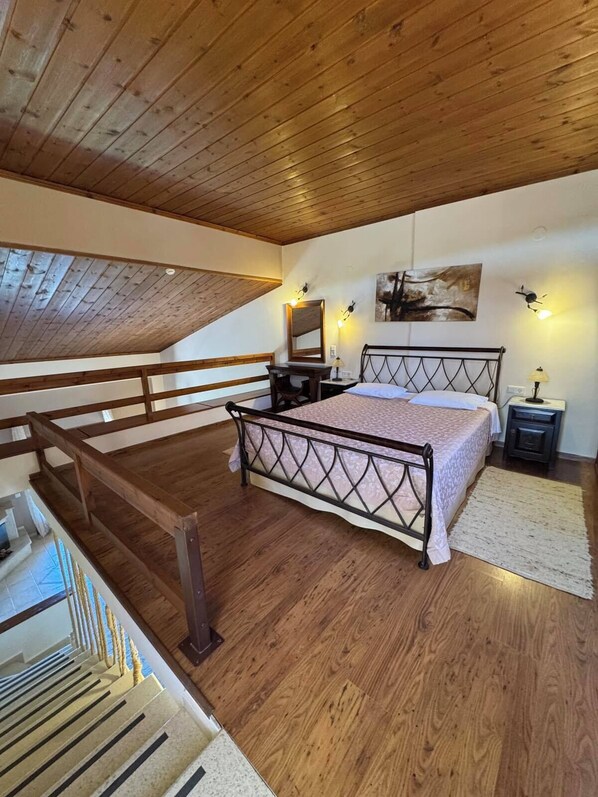 Un dormitorio con techo de madera, cama con estructura de metal, escritorio, silla y un cuadro en la pared.