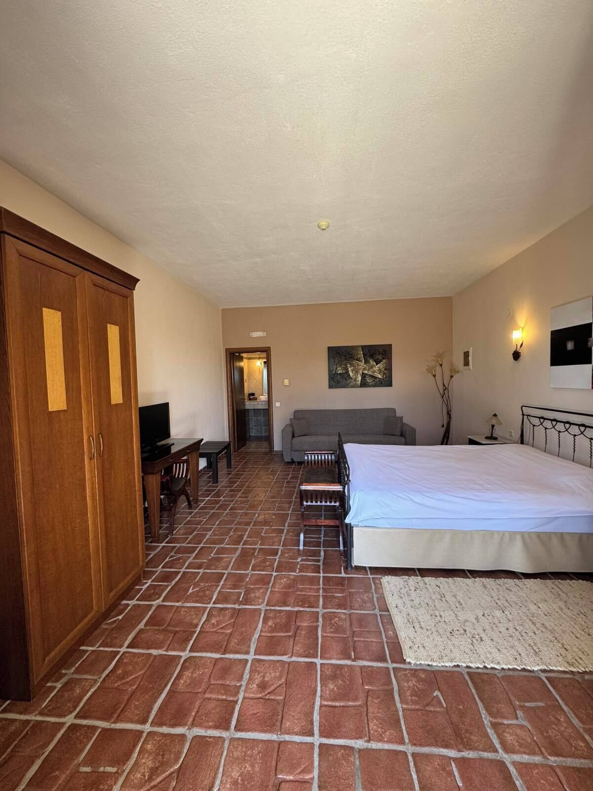 A bedroom with a large bed, a wooden wardrobe, a desk with a chair, a sofa, and a television.