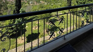 Un balcon doté d’une balustrade en fer forgé, donnant sur un paysage rural avec des arbres et des montagnes à l’horizon.