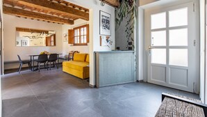 A spacious room with a wooden ceiling, a yellow sofa, a dining area with chairs, a kitchen counter, and a large window.