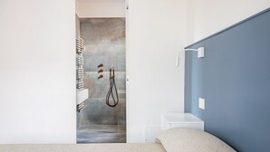 A bedroom with a bed, a white nightstand, a blue wall, and a shower area with a towel rack and showerhead.