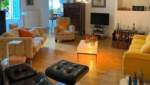 Een ruime woonkamer met een bank, fauteuils, een glazen salontafel en een leren poef. Er is ook een televisie en een kast met decoratieve objecten te zien.