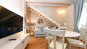 A cozy living room with a TV, sofa, and dining area with chairs and a table. A staircase leads to an upper level.