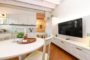 A modern kitchen with white cabinetry, a dining table with chairs, a large flat-screen TV mounted above the counter, and a small potted plant on the counter.