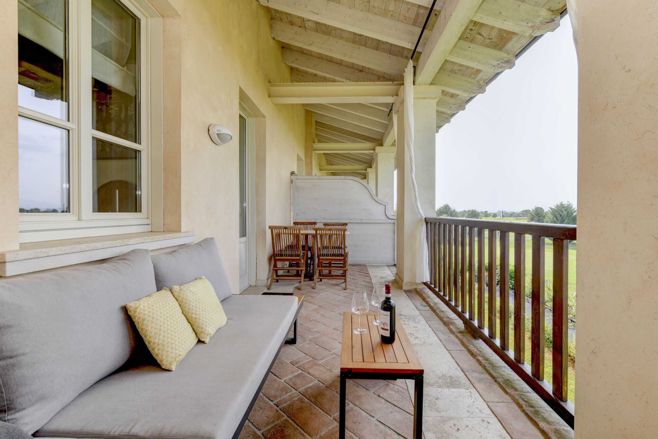 A covered porch with a gray sofa, wooden chairs, and a table with a bottle and glasses.