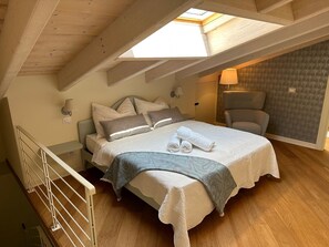 A cozy bedroom with a skylight, a bed with pillows and a blanket, a chair, and a nightstand with a lamp.