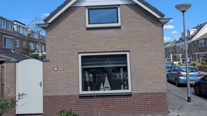 A two-story brick house with a pitched roof, a small window on the upper floor, and a larger window on the ground floor. - The corner house (Egmond aan Zee)