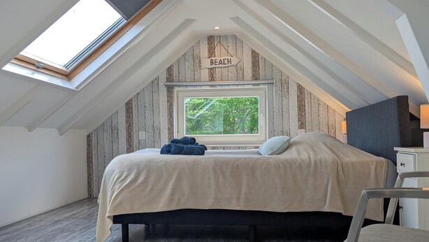 A bedroom with a sloped ceiling, a skylight, a bed, and a window with a view of trees. - The corner house (Egmond aan Zee)