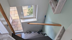 A staircase with a wooden handrail, a small window with a view of the outside, and a white shelf with a vase of flowers. - The corner house (Egmond aan Zee)
