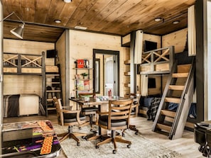 Una habitación acogedora con techo de madera, un escritorio con sillas, una estantería y una cama litera.