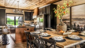 A modern kitchen with dark cabinets, a central island, and a dining area with chairs and a table set for a meal. - Luxury 5BR Getaway | Theater Room | Rooftop Fire Table | Walk to Water Park | Pools | Sleeps 22 (Sevierville)