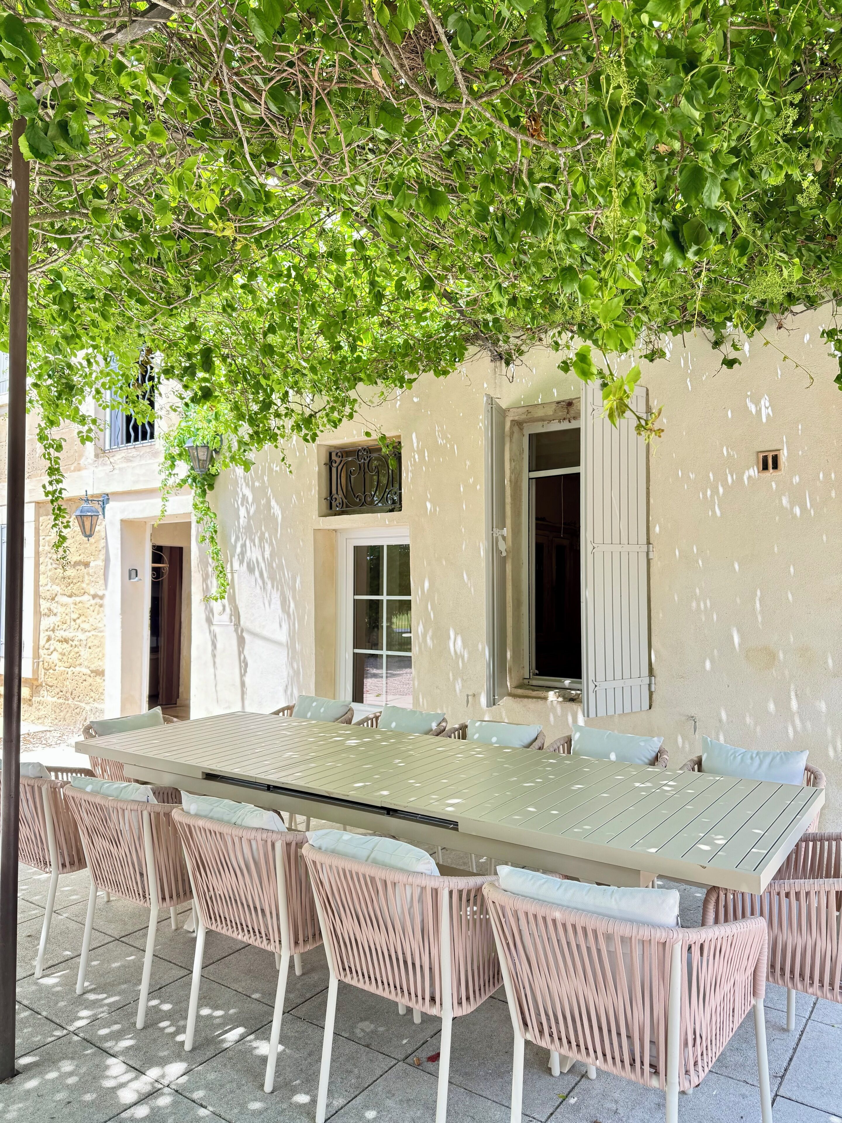 Un área de comedor al aire libre con una mesa larga y sillas de mimbre, rodeada por un edificio con fachada color crema y ventanas.