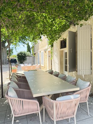 Zona pranzo all'aperto con mobili in vimini e un tavolo grande, immersa nel verde e affiancata da un edificio con finestra e persiana.