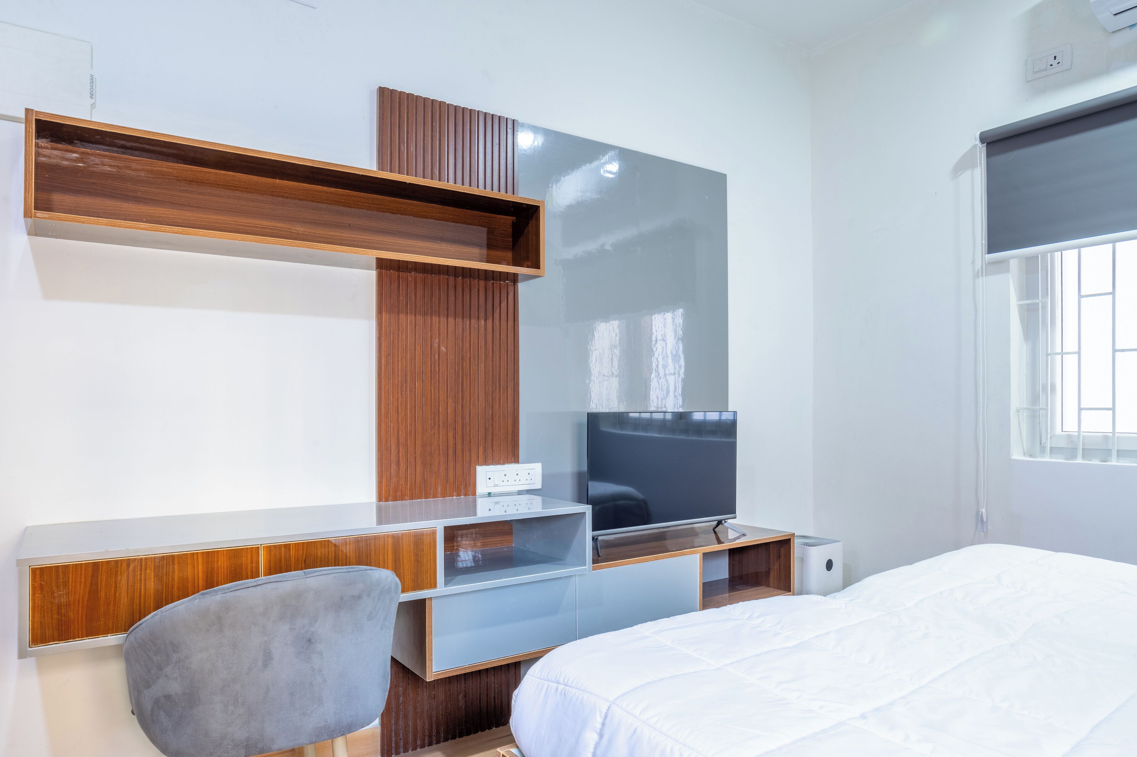 A modern hotel room with a wooden desk, a flat-screen TV, a grey chair, and a large bed.