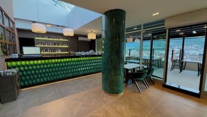 A modern bar area with a green textured wall, a large column, and a view of the city through large windows.