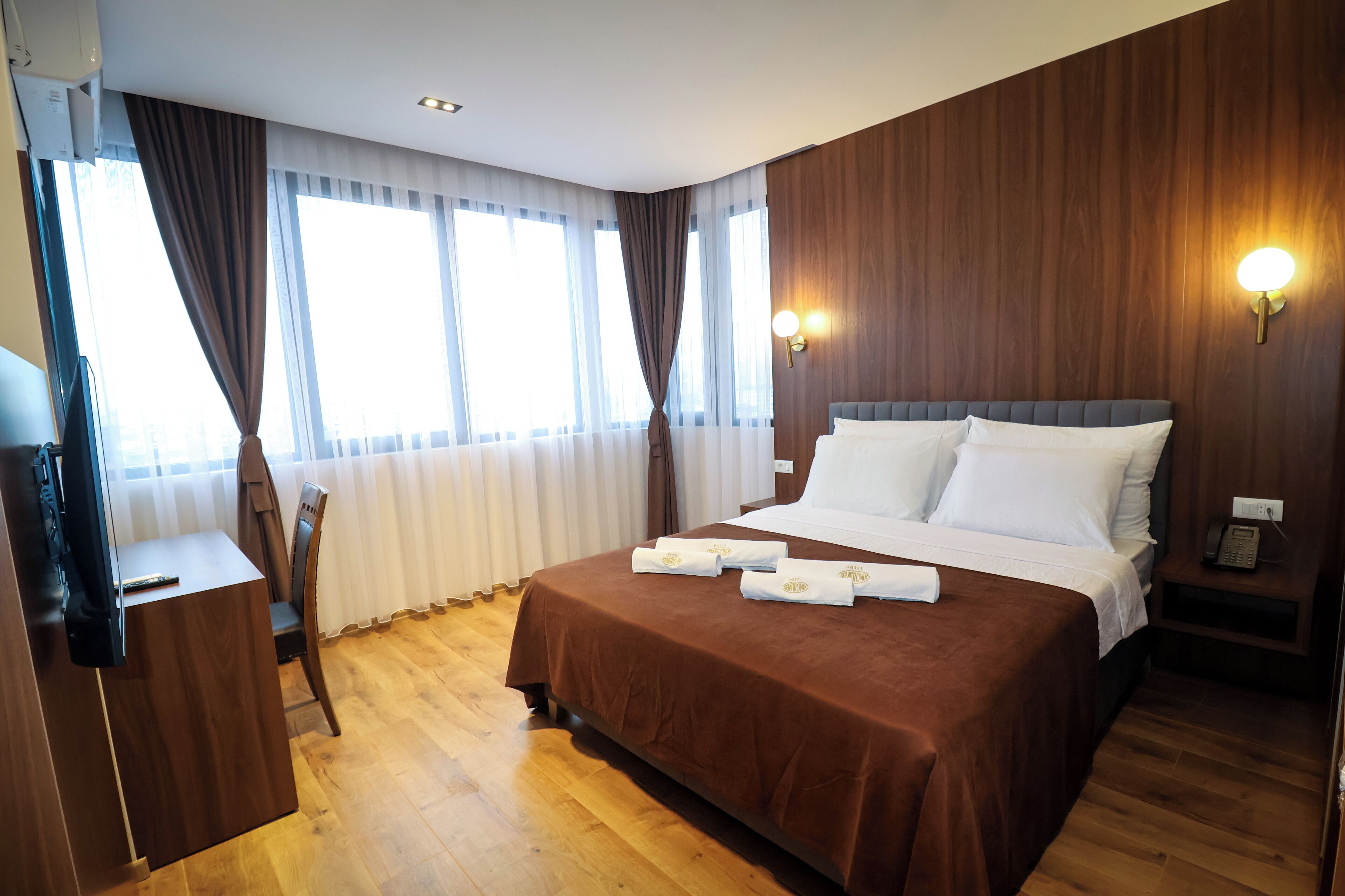A hotel room with a large bed, wooden flooring, a desk with a chair, and a window with curtains.