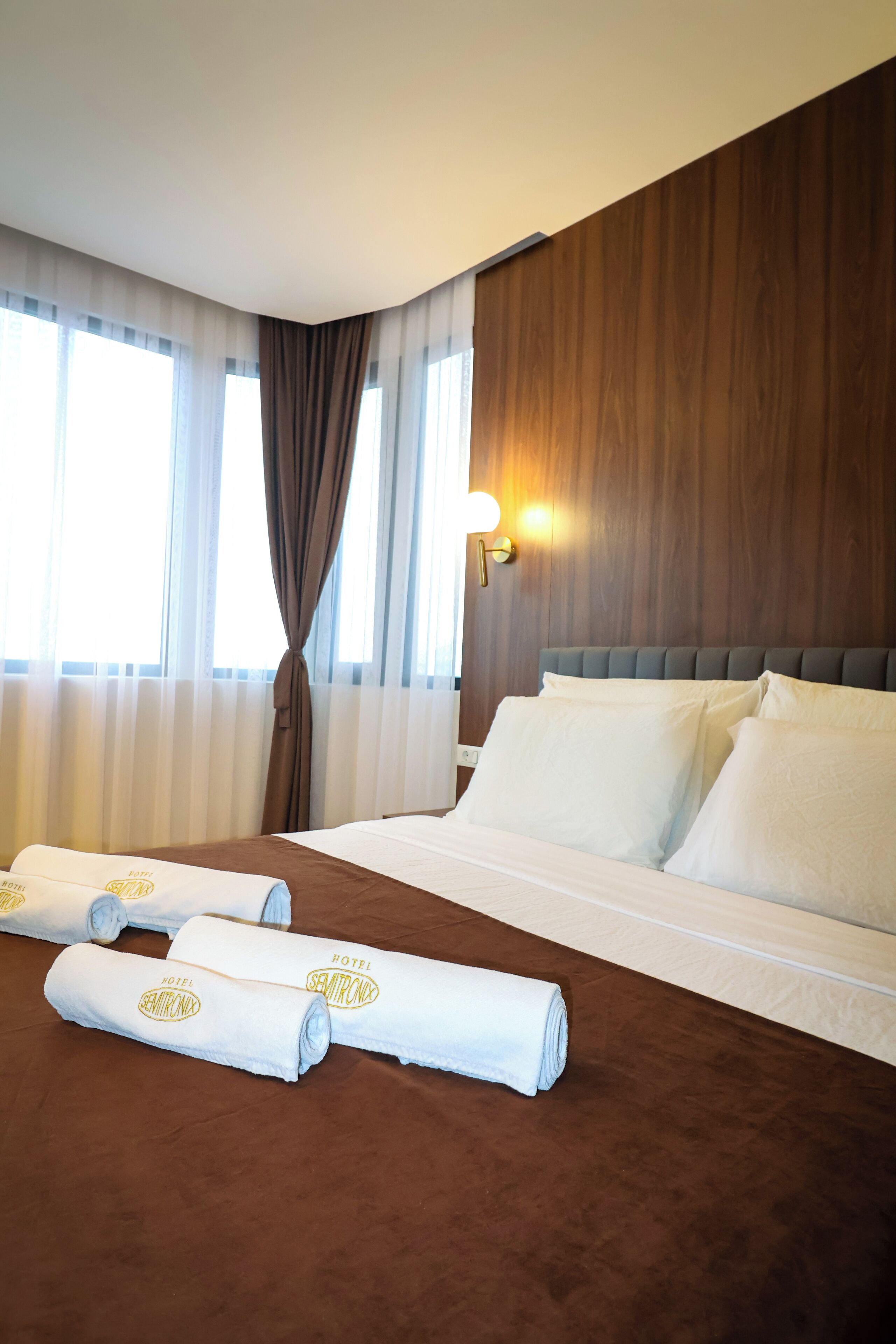 Una camera d'albergo con un letto rifatto, un'ampia finestra con tende e una lampada a parete.
