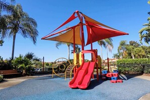 Ein Spielplatz mit einer roten Rutsche und Klettergerüsten, umgeben von Palmen und blauem Himmel.