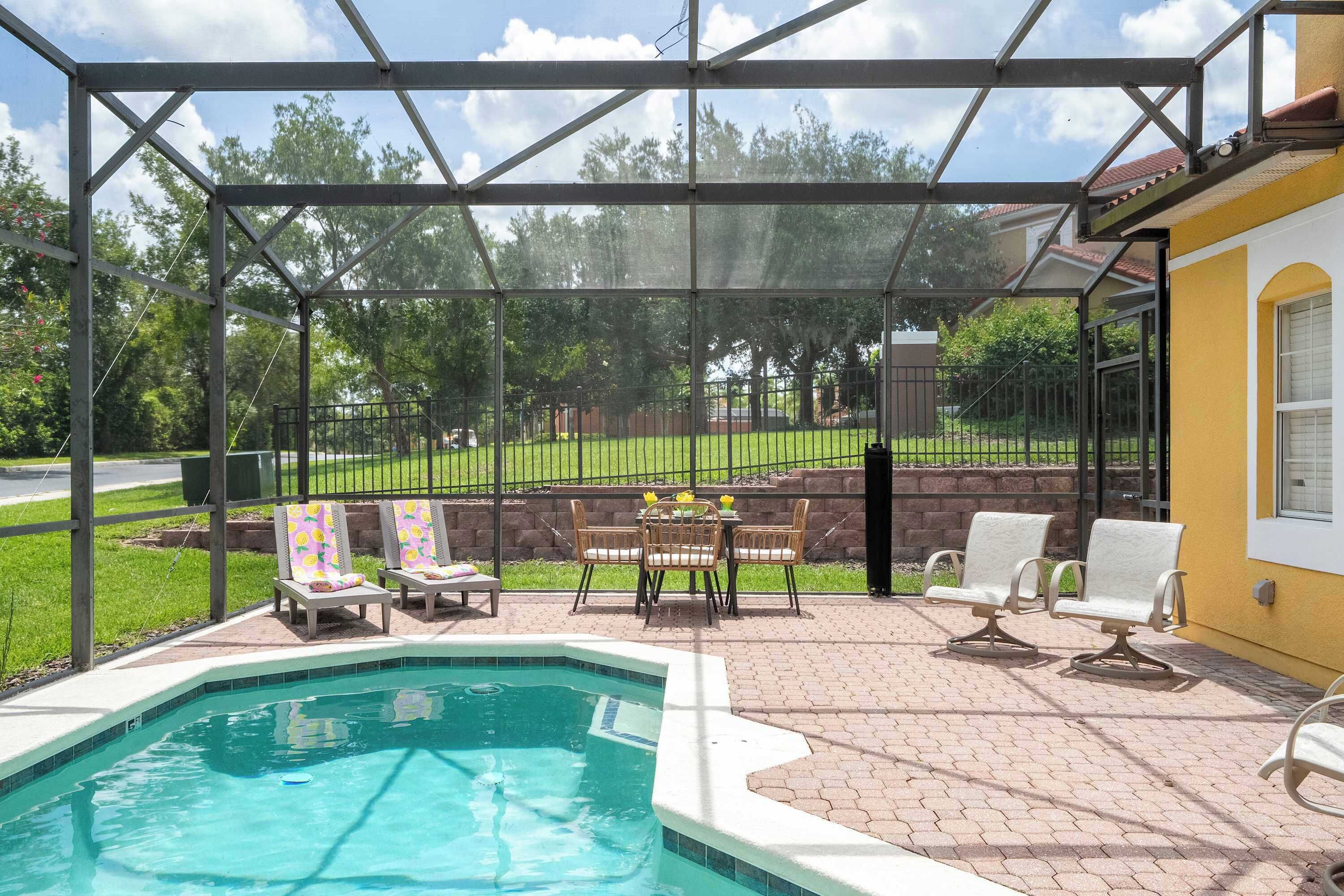 A covered outdoor swimming pool with a patio area, a table with chairs, and a view of a green landscape.