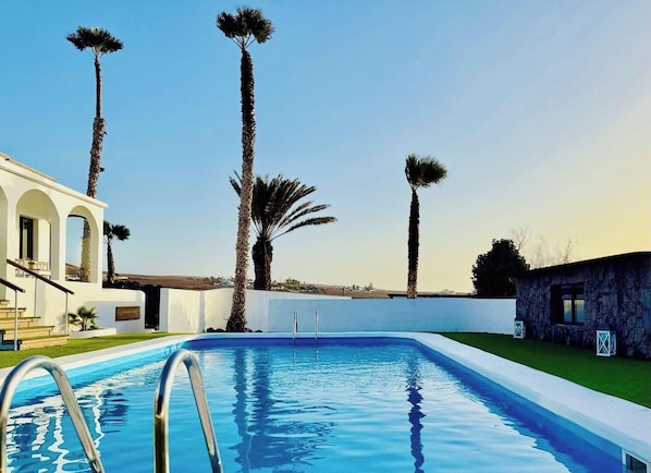 A swimming pool with a clear blue water surface, surrounded by palm trees and a white building with arches.