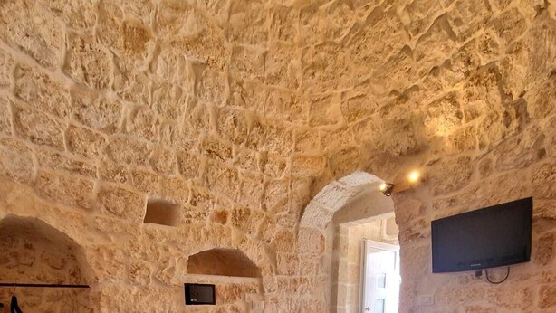 A stone-walled room with a bed, a flat-screen TV, and a small wooden chest.