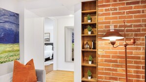 Een moderne woonkamer met een grijze bank, een groot abstract schilderij en een bakstenen muur met een houten plank waarop decoratieve objecten en een vloerlamp staan.
