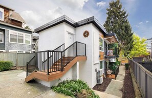 A residential building with a wooden staircase leading to a white door, surrounded by a garden and a wooden fence.