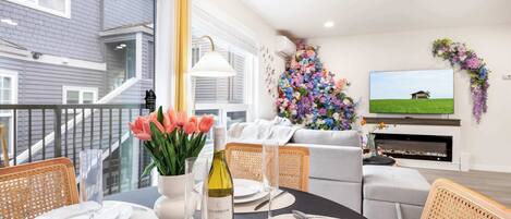A dining area with a black table set for four, white plates, and a bottle of wine. There is a large floral arrangement on the wall and a television mounted above a fireplace.