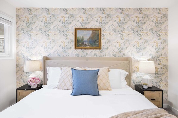 Un lit bien fait, avec un oreiller bleu, du linge de lit blanc et un papier peint à motifs floraux. On y trouve deux tables de chevet avec des lampes et un tableau encadré au-dessus du lit.