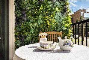 A table with a white teapot and cup set on a white tablecloth, placed on a balcony with a green plant wall in the background.